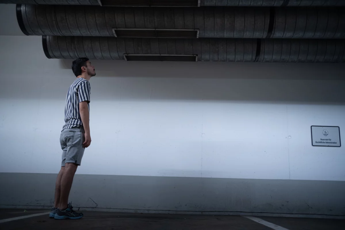 Illuminated underground parking garage. A single performer stands at the left side of the frame and looks at the ceiling.