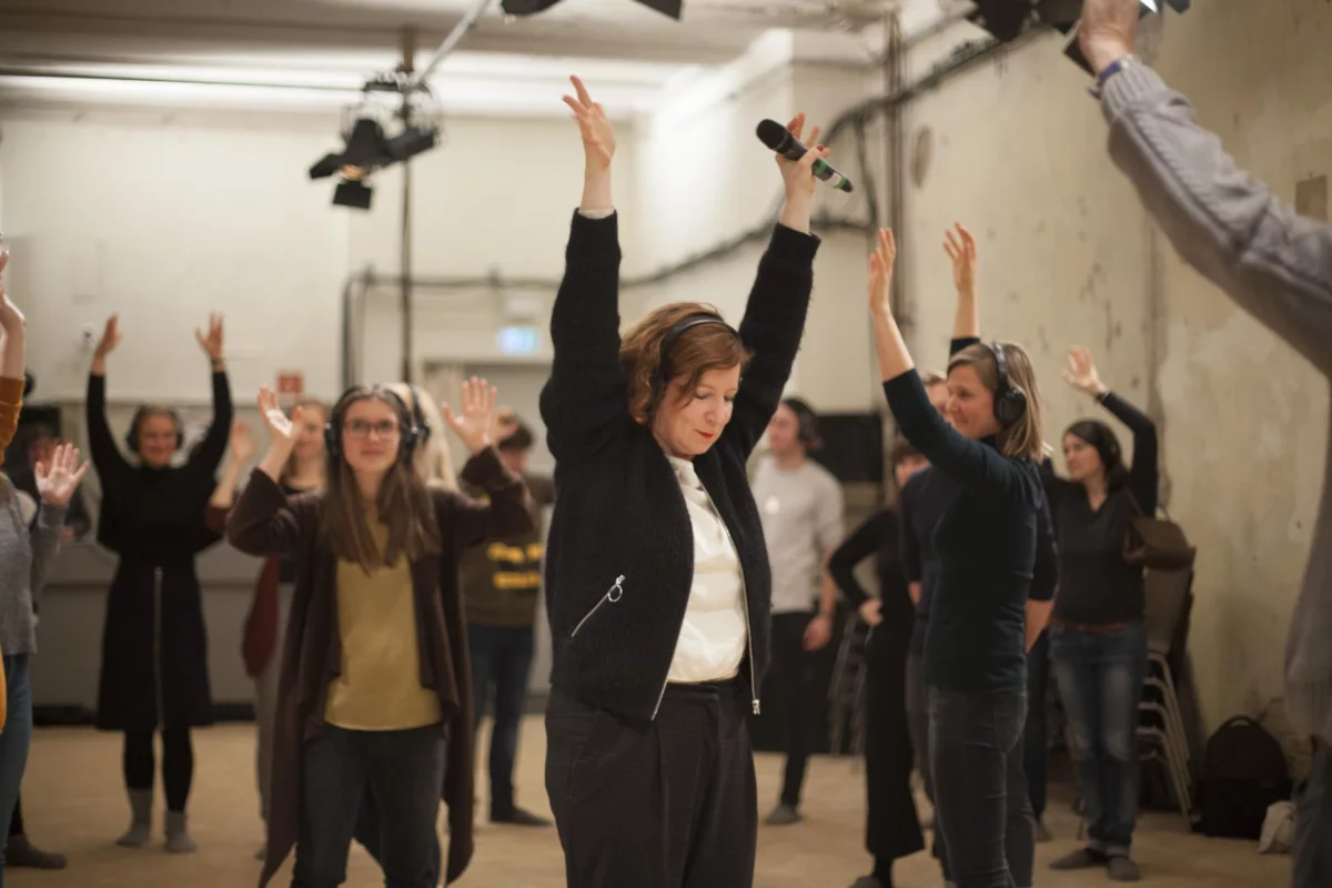Im Zentrum steht eine Performerin. Sie trägt Kopfhörer, streckt die Arme in die Luft und hat ein Mikrofon in der Hand. Um sie herum steht das Publikum, das auch die Arme in die Luft streckt und Kopfhörer trägt.