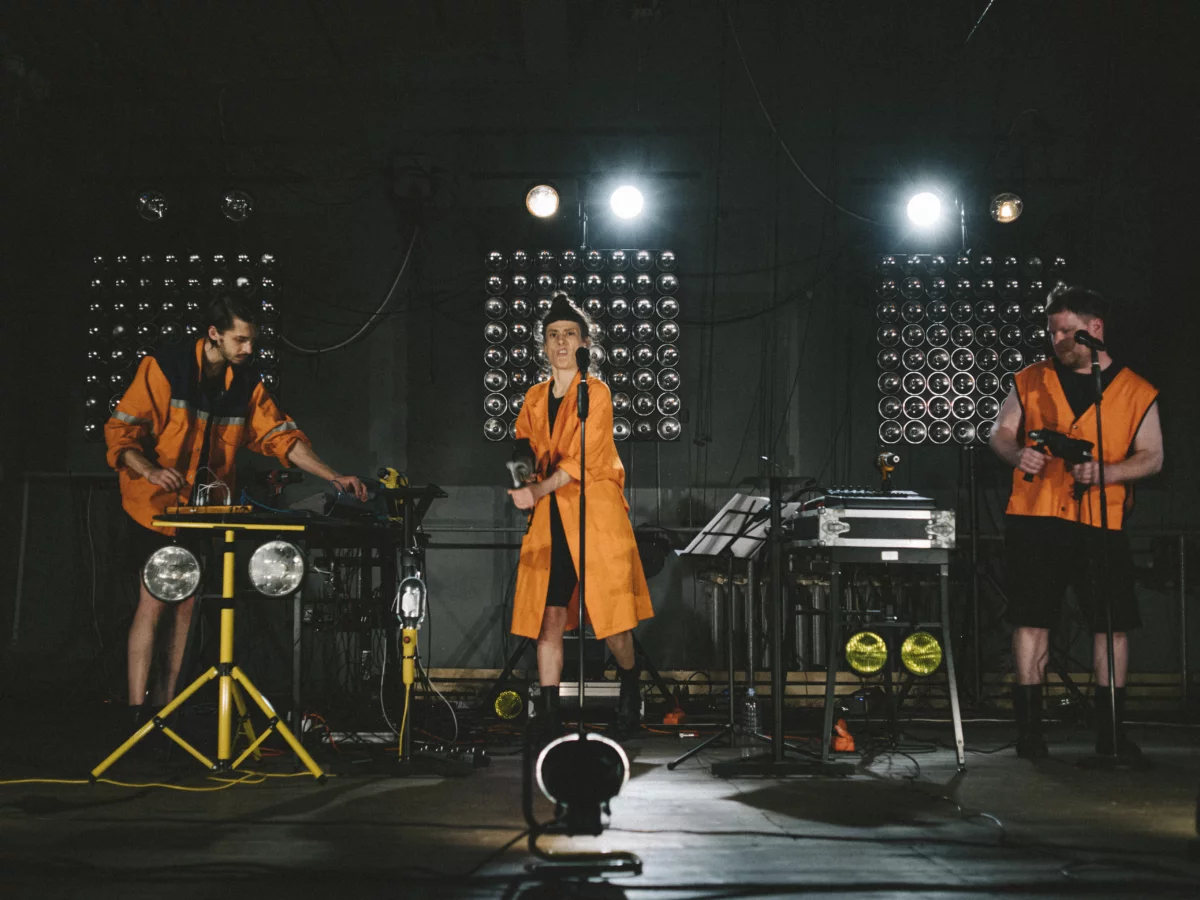 Szene aus einer Performance von Teatru Spalatorie: Drei Performer*innen auf der Bühne in Arbeiter*innen-Outfits. Ein Performer bedient ein Technikpult, die beiden anderen halten Bohrmaschinen, die sie wie Instrumente behandeln.