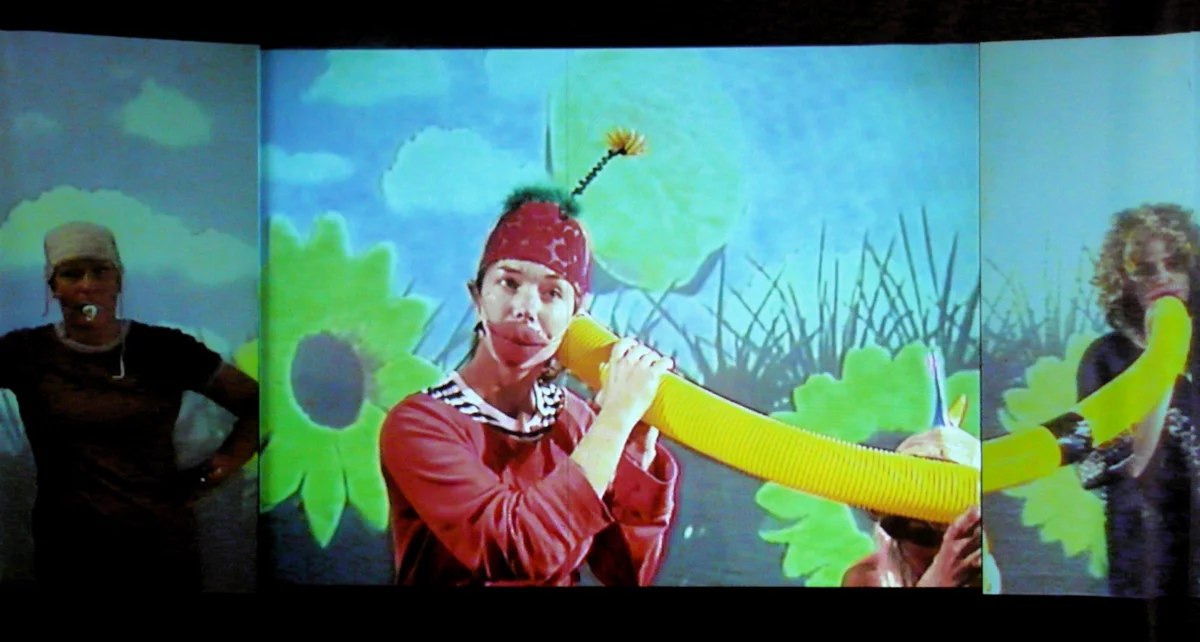 The projection of a flower meadow, in front of it three performers: One in penumbra, one wearing oversized red cloth lips and a cap with green cloth and a flower attached to the top. The person holds the end of a tube to her ear. The third performer holds the other end of the tube in front of her mouth.
