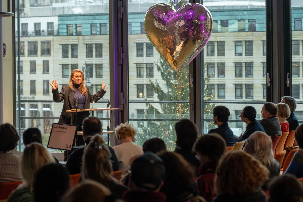 Jagoda Marinic hält ihren Vortrag vor einem voll besetzten Auditorium. Neben ihr schwebt ein goldenes Herz. Durch die Fenster im Hintergrund sind die Häuser des Pariser Platzes in Berlin zu erknennen.