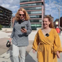 Zwei Personen bewegen sich im Stadtraum. Eine Person schaut während des Gehens auf ihr Handy.