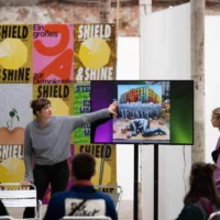 Two people are standing to the left and right of a large screen showing a performance scene: a man in a silver metallic suit is lying on a piece of cardboard in an embryonic pose. Behind him is a picture of a crowd of people dressed in grey demonstrating in front of a colorful house.