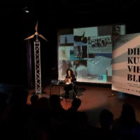The artist and art professor Isabel Lewis sits on the dimly lit stage and gives her lecture "On hospitality and the body". Images of performance situations from her lecture are projected in the background.