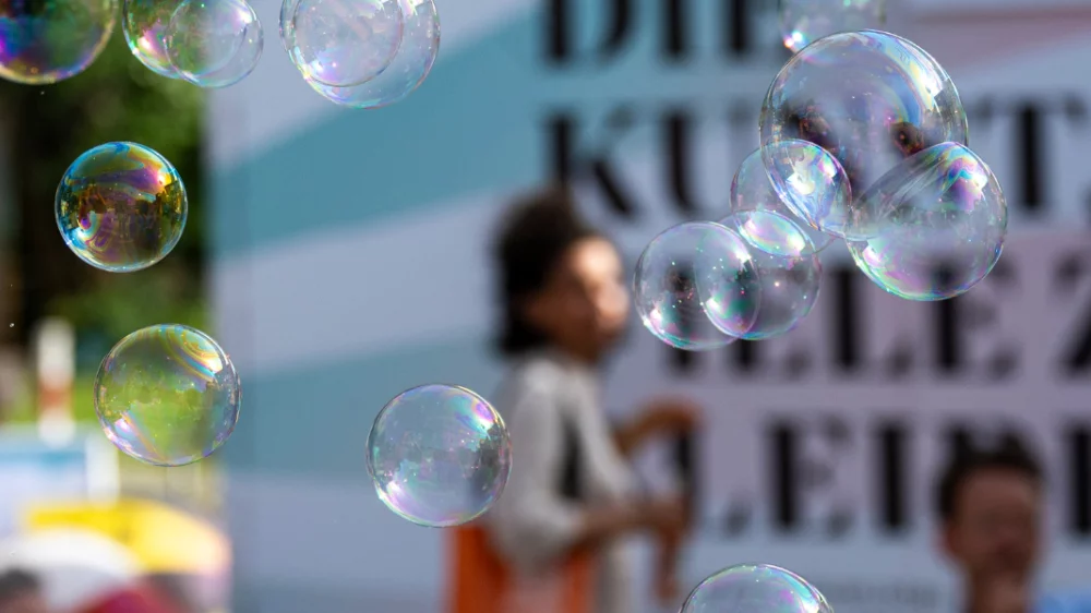 Soap bubbles fly through the air. Blurred in the background, people can be seen talking in front of a billboard with the inscription “DIE KUNST, VIELE ZU BLEIBEN” (“THE ART OF BEING MANY”) in a summery setting.