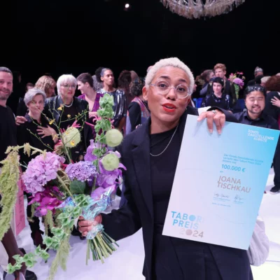 Choreographer Joana Tischkau holds the Tabori Prize certificate and a bouquet of flowers up to the camera with a smile and is delighted with the award. The audience at the award ceremony can be seen in the background.