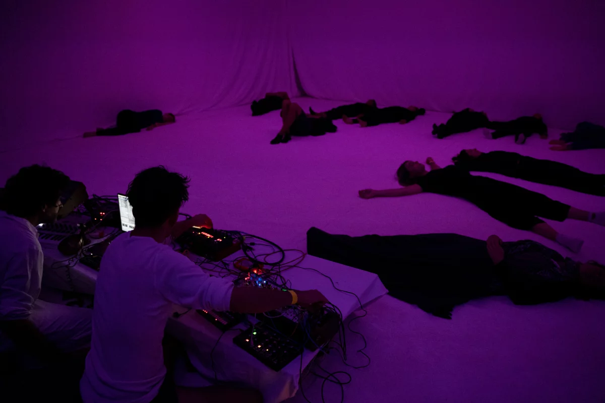 A purple illuminated stage. Two people are sitting at a technical console. Eleven people are lying on the floor in front of you.