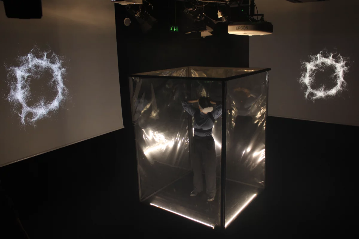 Eine Person mit VR-Brille steht in einem durchsichtigen hohen Kubus, der in einer dunklen Bühnensituation installiert ist. Rechts und links davon finden sich Leinwände auf die projiziert wird.