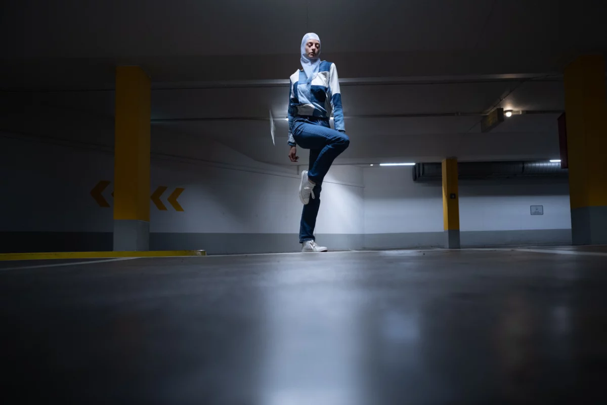 Dark underground parking lot with a light spotting on a dancer wearing a headscarf and standing gracefully on one leg.