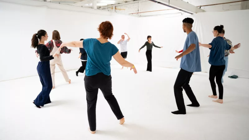 A room with white walls and white floor. Nine people stand in a circle facing each other and dance.