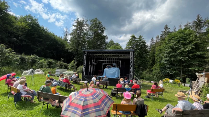 An outdoor performance of the puppet theater. A play is shown on a black stage. In front of it, the audience sits on the lawn with benches and umbrellas.