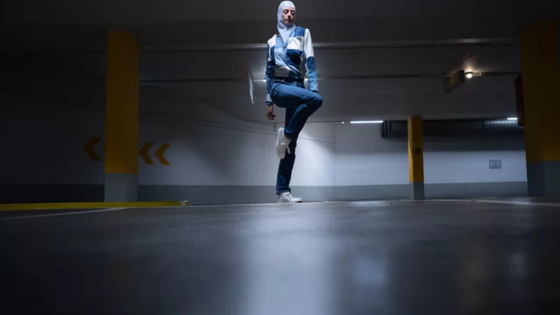 Dark underground parking lot with a light spotting on a dancer wearing a headscarf and standing gracefully on one leg.