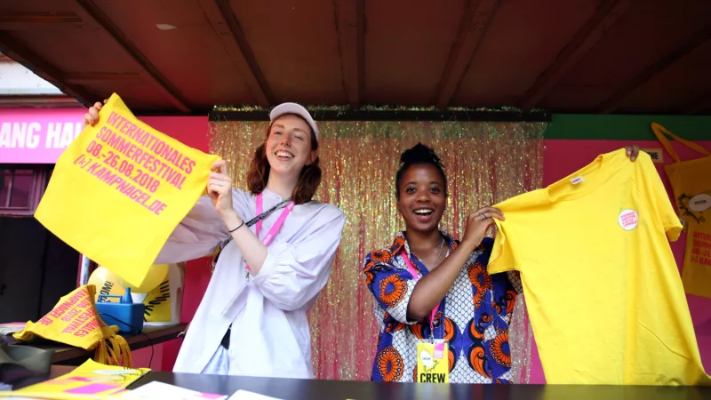 Zwei junge Personen stehen an einem Counter und halten ein gelbes T-Shirt und einen gelben Beutel mit dem Aufdruck vom Internationalen Sommerfestivals von Kampnagel in die Kamera.