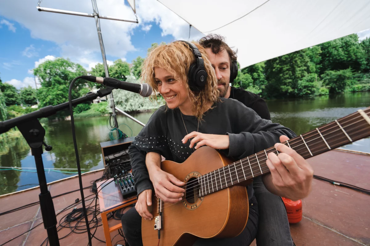 A covered footbridge, on a river. Two musicians sit behind each other and play a guitar together with four hands. Both are wearing headphones.