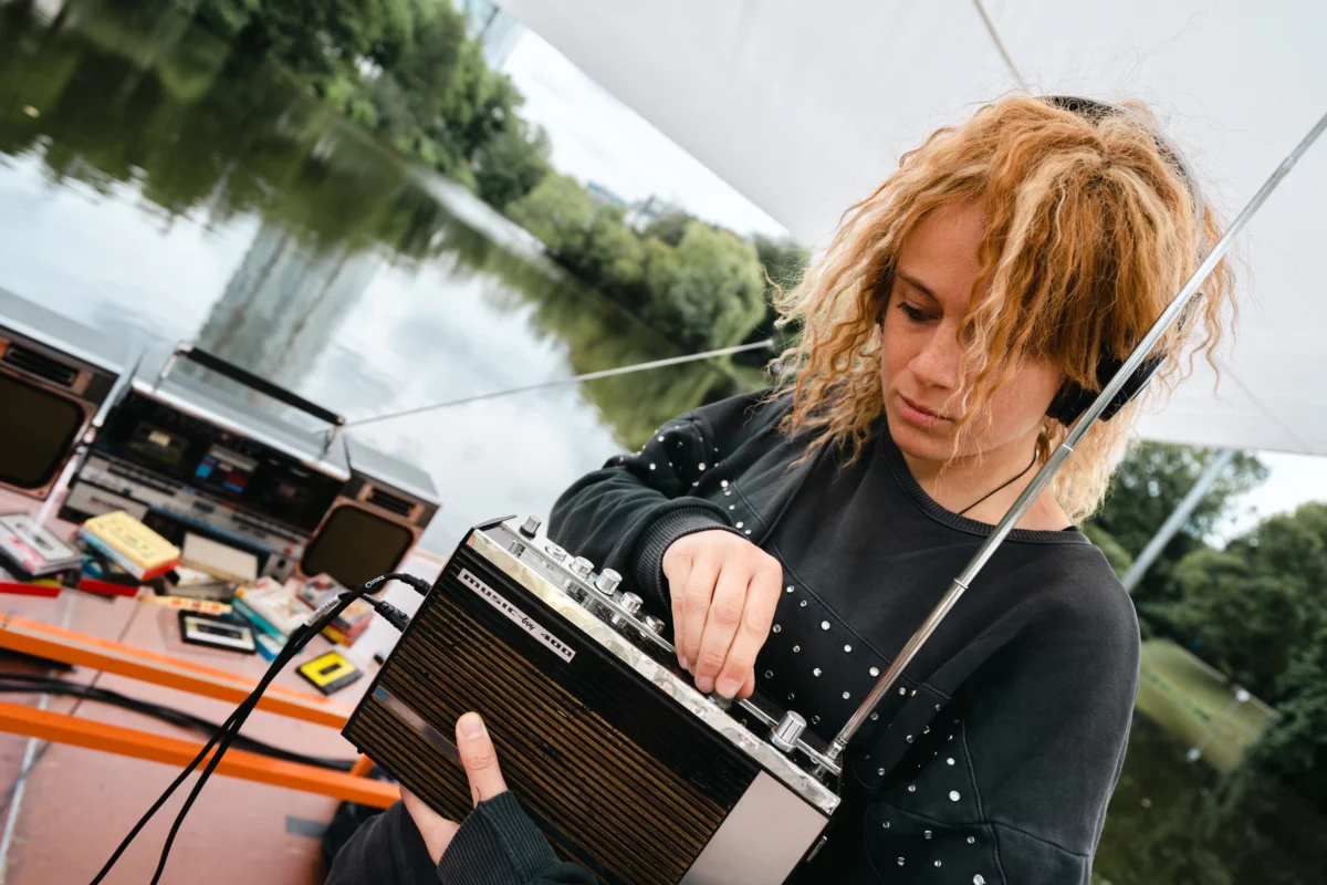 A covered footbridge, on a river. A performer holds a portable radio in her arms and turns the dial. Behind her, on a table, stands a large cassette radio, in front of which lie many cassettes and cassette sleeves.