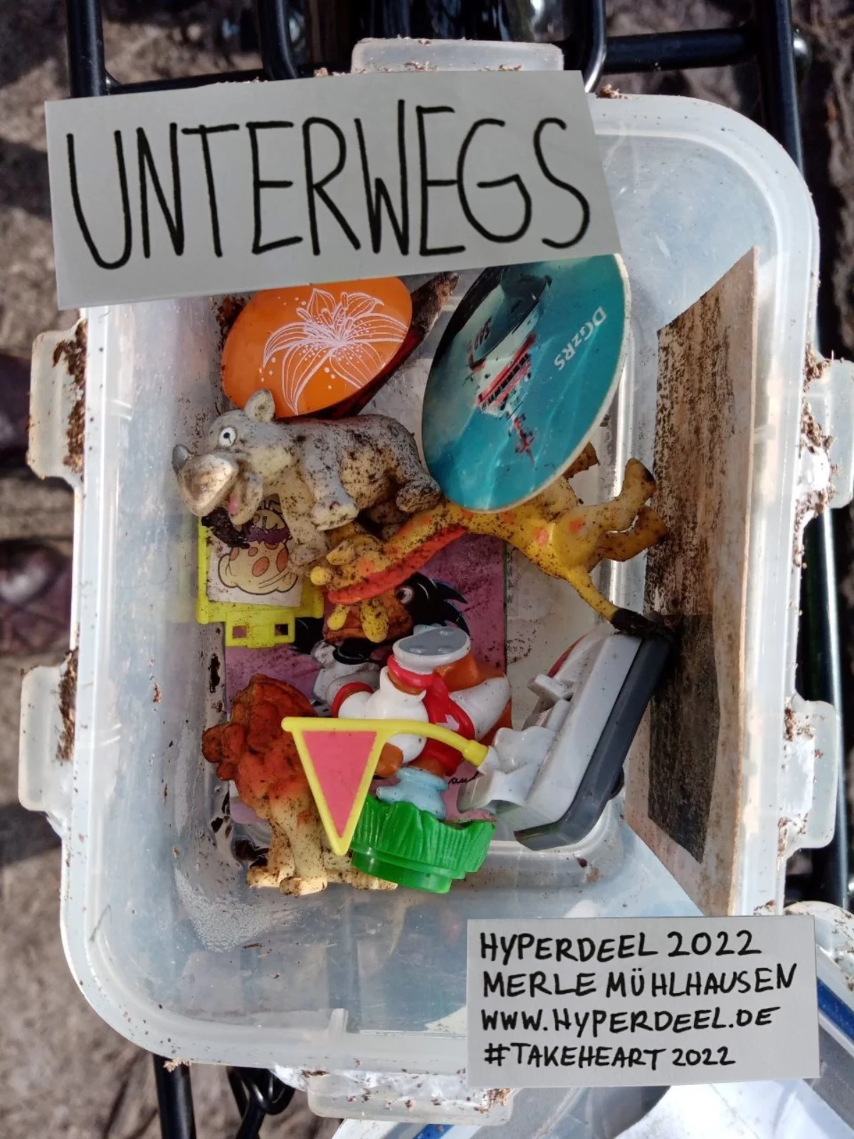 View into a plastic box in which there are old, sandy toy figures. At the bos are two signs on one says: On the road. The other says: Hyperdeel 2022, Merle Mühlhausen, www.hyperdeel.de, #TakeHeart 2022.