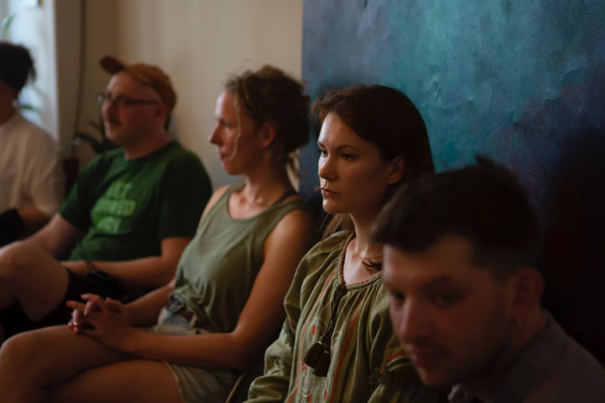 Five people are sitting on chairs in a row against a wall. Behind them hangs a picture. You see them diagonally from the side, they look dreamily and do not talk.