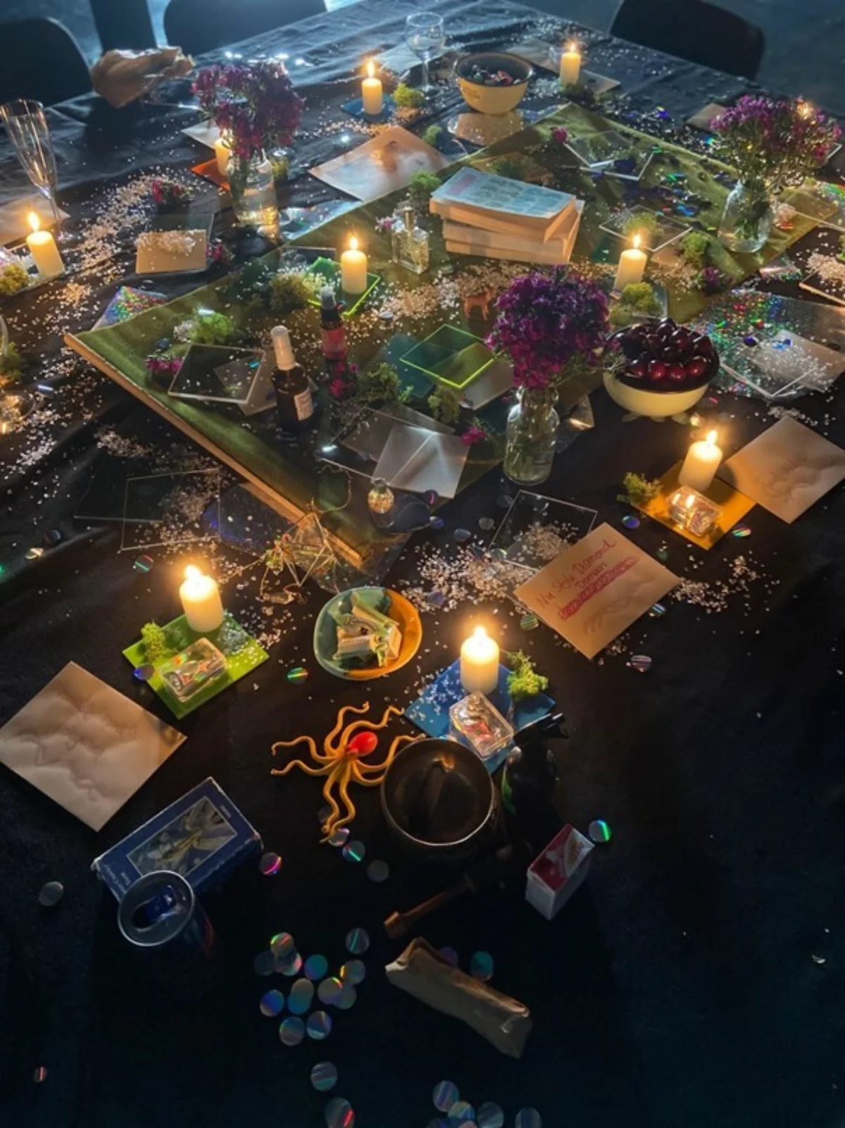 Ein Tisch auf dem eine rituelle Séance hergerichtet ist. Im Zentrum ein Tuch von dem ausgehend Bücher, Schalen mit Lebensmitteln, Kerzen und Gläser arrangiert sind.