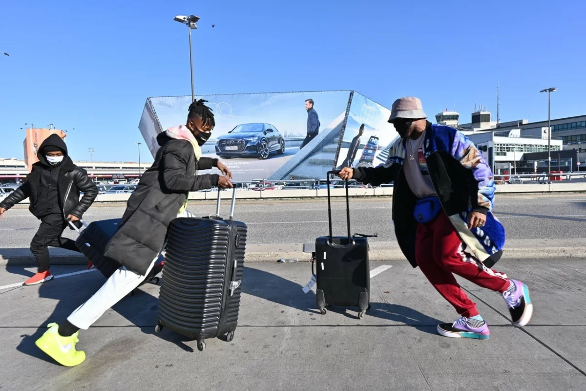 Parkplatz am Flughafen Tegel. Drei Tänzer*innen tragen medizinische Masken und rennen, mit jeweils einem schwarzen Rollkoffer umeinander herum.