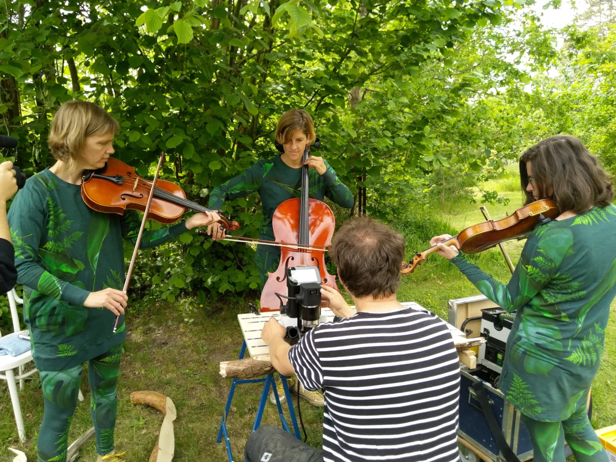 Ein Park. Drei Personen tragen Kleidung, die mit Blättern bedruckt ist. Zwei spielen Geige, eine Violine. Alle drei sind einem Tisch zugewandt an dem eine weitere Person sitzt.
