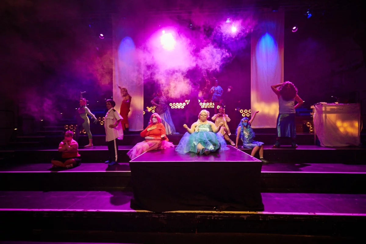 Scene from "The Ball": A step-like stage situation with a centered footbridge projecting forward. The stage is bathed in fog and pink light. The performers of Meine Damen and Herren sit and stand on the catwalk and steps in colorful costumes, dresses and wigs.