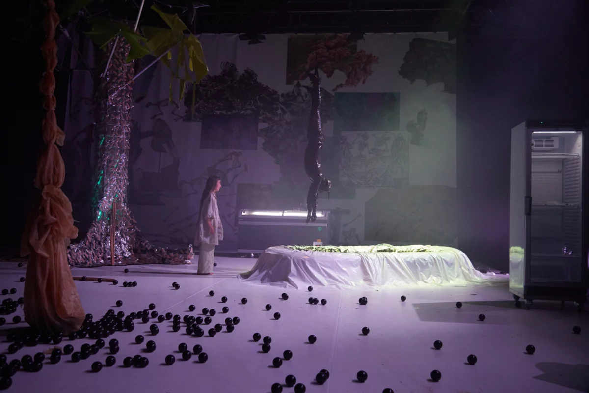 Scene from "World without us": A white stage floor on which a plunge pool stands. Above it hangs a person, suspended upside down by his feet, he has just been pulled out of the basin and is completely covered with black paint. A performer stands in front of it and watches the scenario.