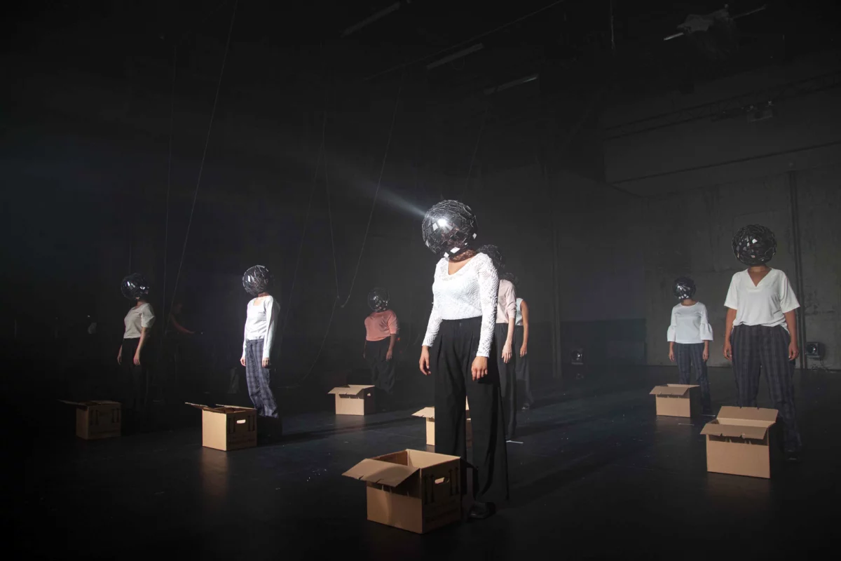 Eight performers stand in three rows on a dark stage. Each of them has a cardboard moving box in front of them and a disco ball over their head, which is reflected by the light.