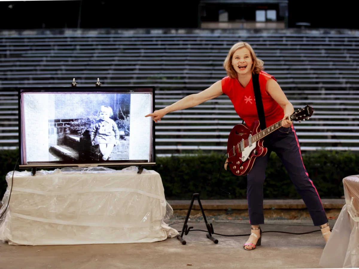Bernadette La Hengst has an electric guitar strapped around her and points to a screen standing next to her. On it is a black and white photograph with a small child.