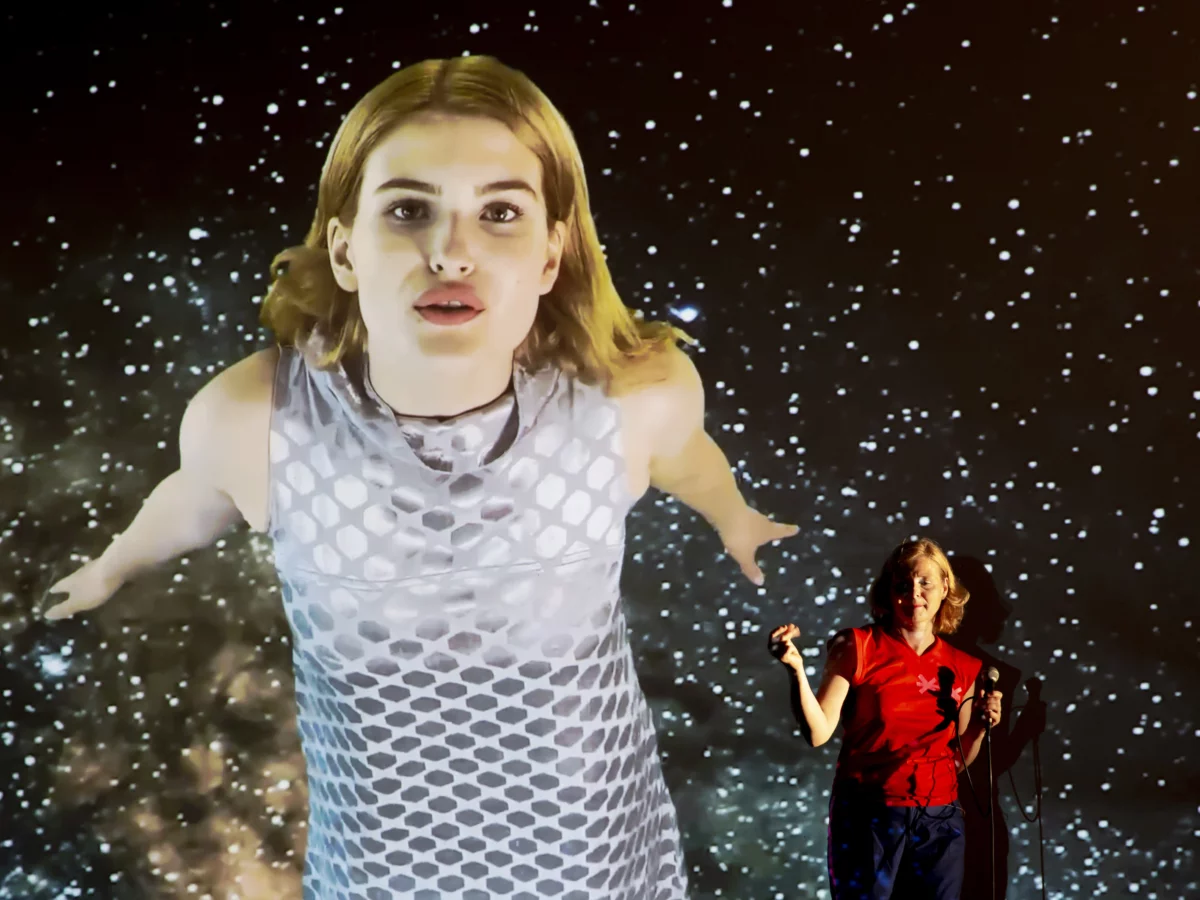 Bernadette La Hengst tanzt vor einer Leinwand, auf die eine junge Frau projiziert ist. In ihrem Hintergrund ein Sternenhimmel.