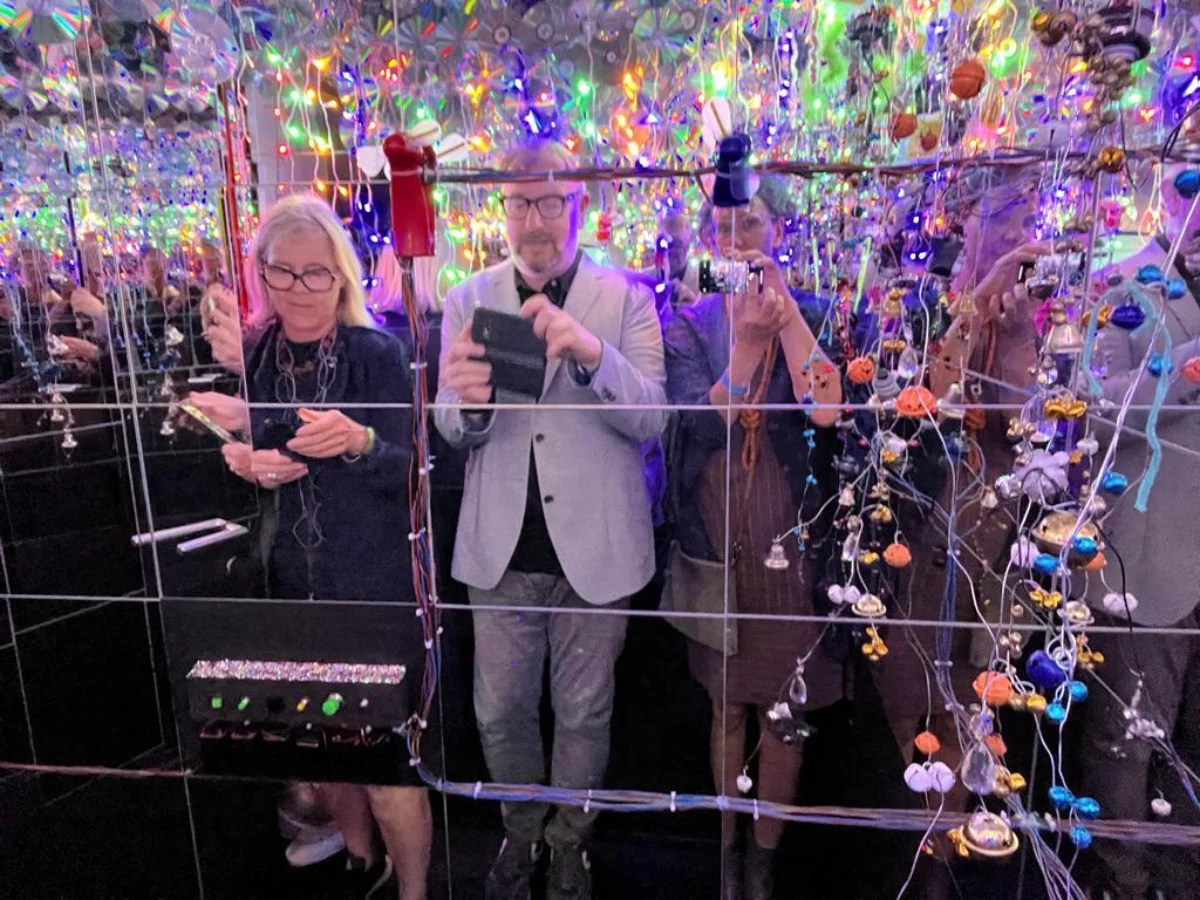 View into a distorting mirror. Bettina Milz, Holger Bergmann and a third person are visible. They are all looking at the displays of the cell phones in their hands. Behind them are many colorful strings of lights. On the mirror itself are some cables and buttons and colorful garlands.