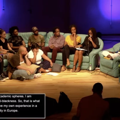 10 people on a sofa in a studio, one person speaks into a microphone, the audience sits in front. In the corner of the picture a field in which live translation is running.