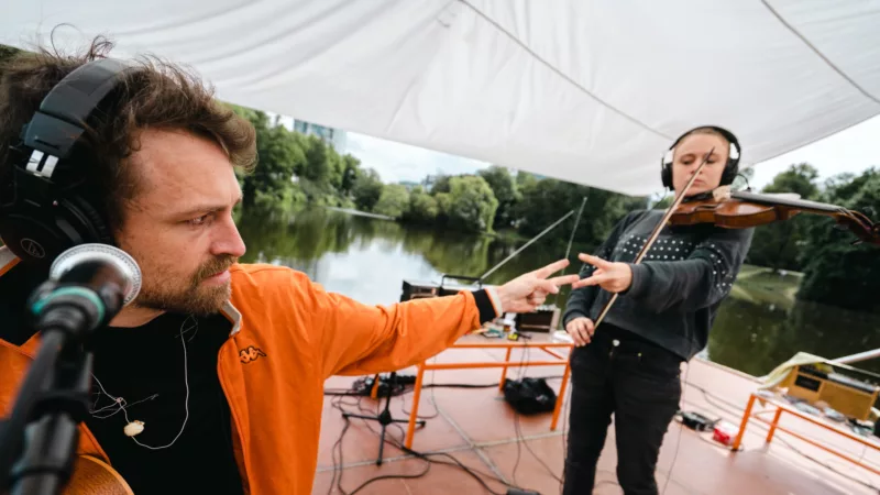 A covered footbridge, on a river. On it two musicians. One person at the microphone with headphones, one person with headphones and violin. Both are facing each other and playing paper scissors.