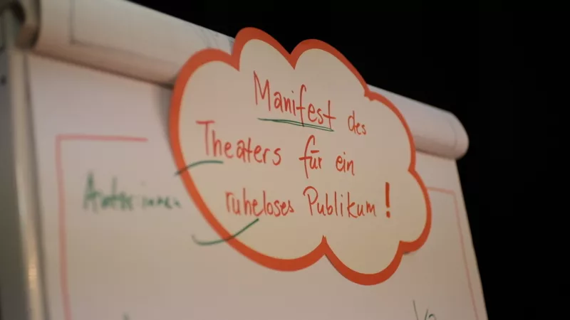 A flipchart with a moderation card stuck to the top. On it you can read "Manifesto of theater for a restless audience".