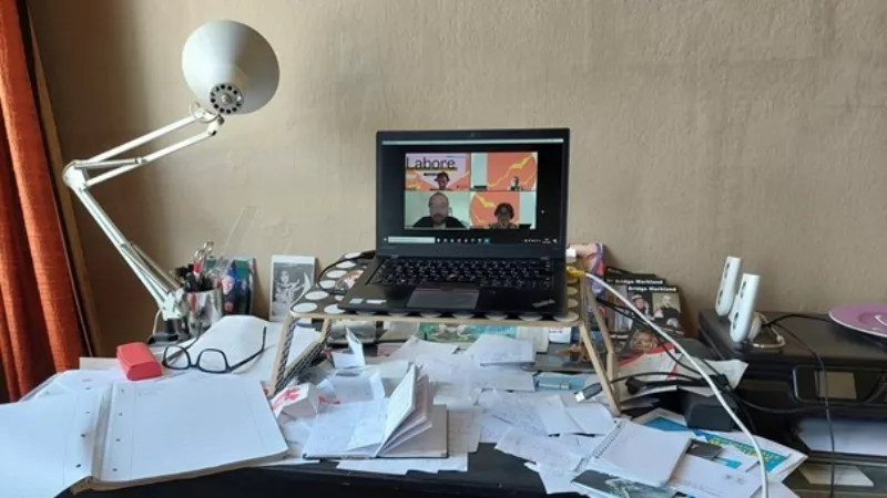A desk. Covered all over with paper, notepads and similar screaming accessories. Somewhat elevated is a laptop on which an online conference with three other people is taking place.
