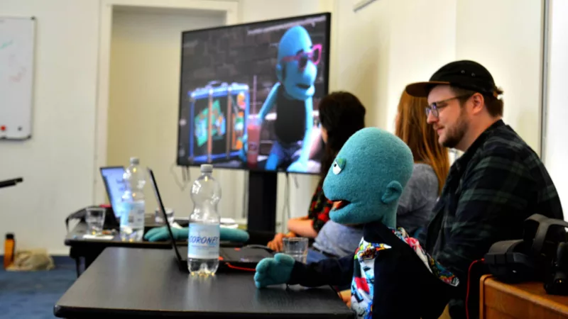Konferenzraum. Zwei Tische im Bild, daran drei Personen und eine Puppe. Im Hintergrund ein Bildschirm, mit einem Film auf dem die Puppe zu sehen ist.