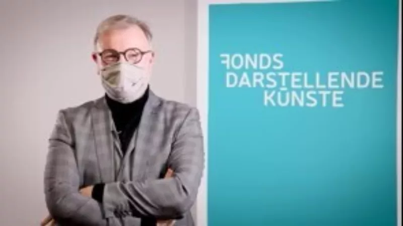 Holger Bergmann trägt einen grau karierten Anzug, hat graues Haar. Er hat die Arme vor dem Körper verschränkt, trägt eine Brille und einen grauen Mund-Nasenschutz. Im Hintergrund steht ein türkises Roll-up mit der Aufschrift Fonds Darstellende Künste.