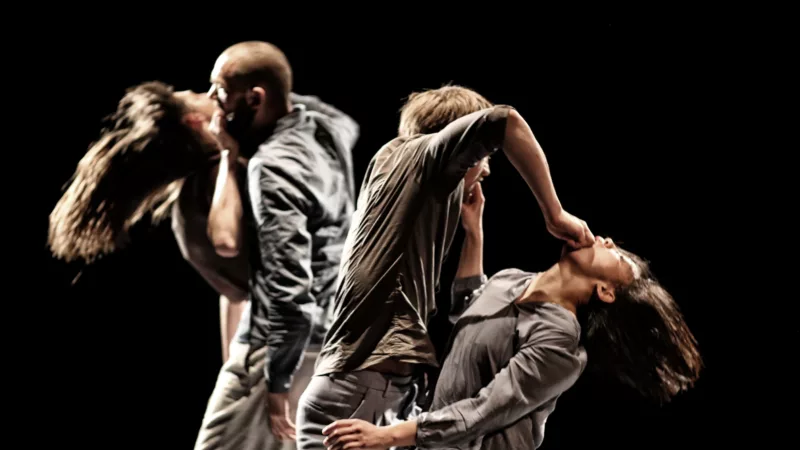Two performers are in one movement. They each have their own hand in their partner's mouth. One person's head is bent backwards, the other bends slightly above him. In the background a similarly arranged couple can be seen.