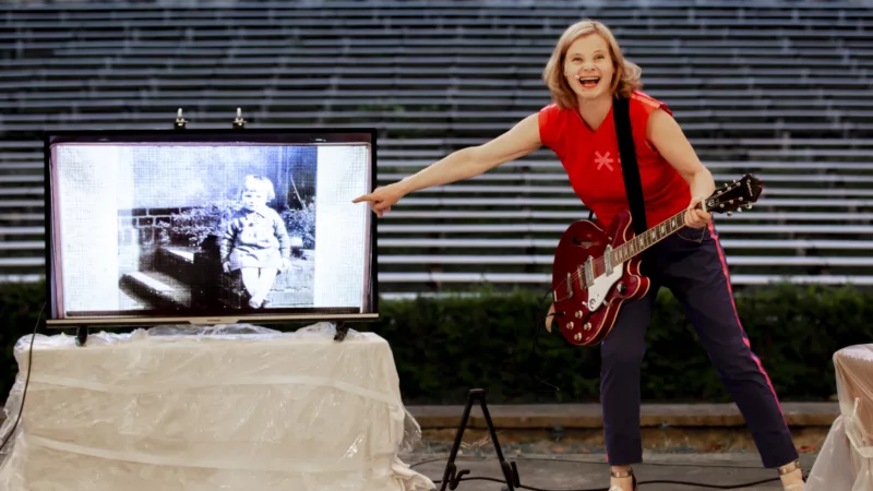 Bernadette La Hengst has an electric guitar strapped around her and points to a screen standing next to her. On it is a black and white photograph with a small child.