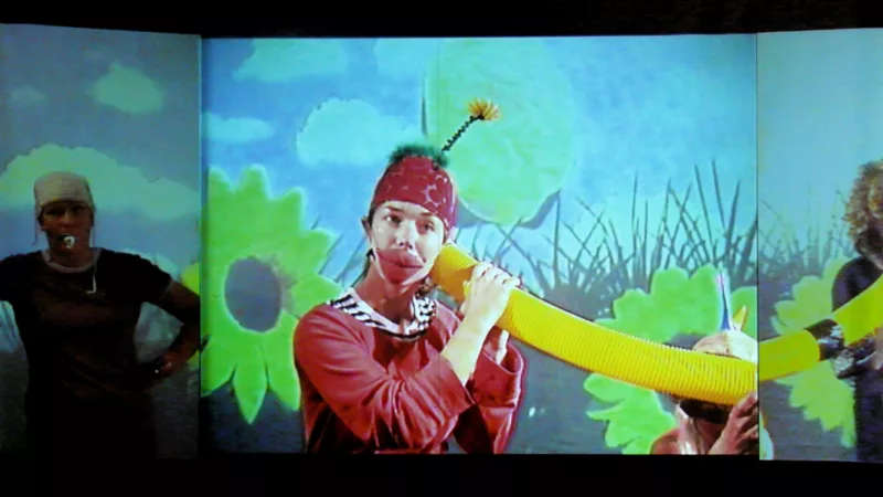 The projection of a flower meadow, in front of it three performers: One in penumbra, one wearing oversized red cloth lips and a cap with green cloth and a flower attached to the top. The person holds the end of a tube to her ear. The third performer holds the other end of the tube in front of her mouth.