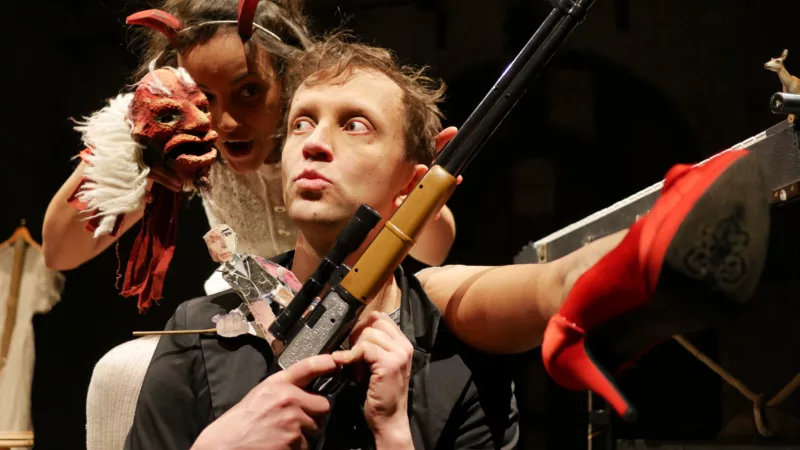 A performer sits, holding a plastic gun in front of him and a paper doll. Behind him is a female performer who puts her leg over his shoulder in high heels. She wears devil horns on her head and holds a hand puppet close to his head.
