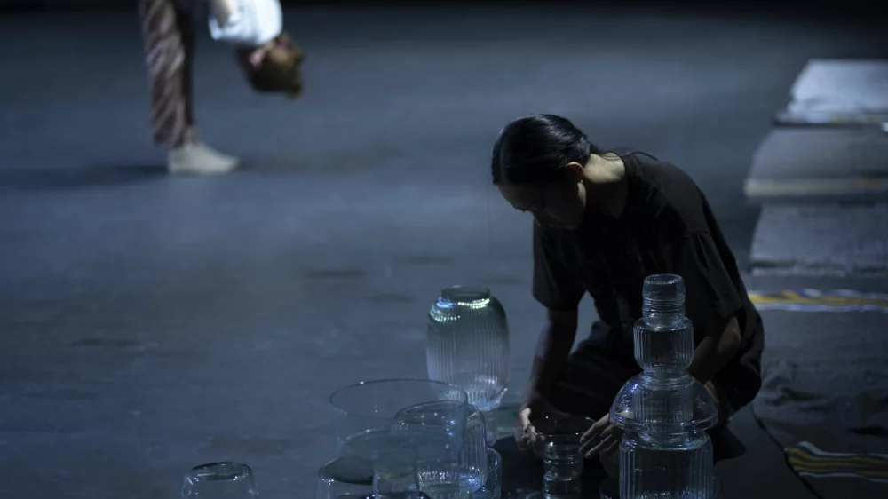 Stage floor. In the background stands a performer who bends her upper body to her feet. In the foreground, a performer kneels in front of glasses and glass vases, some of which are stacked.