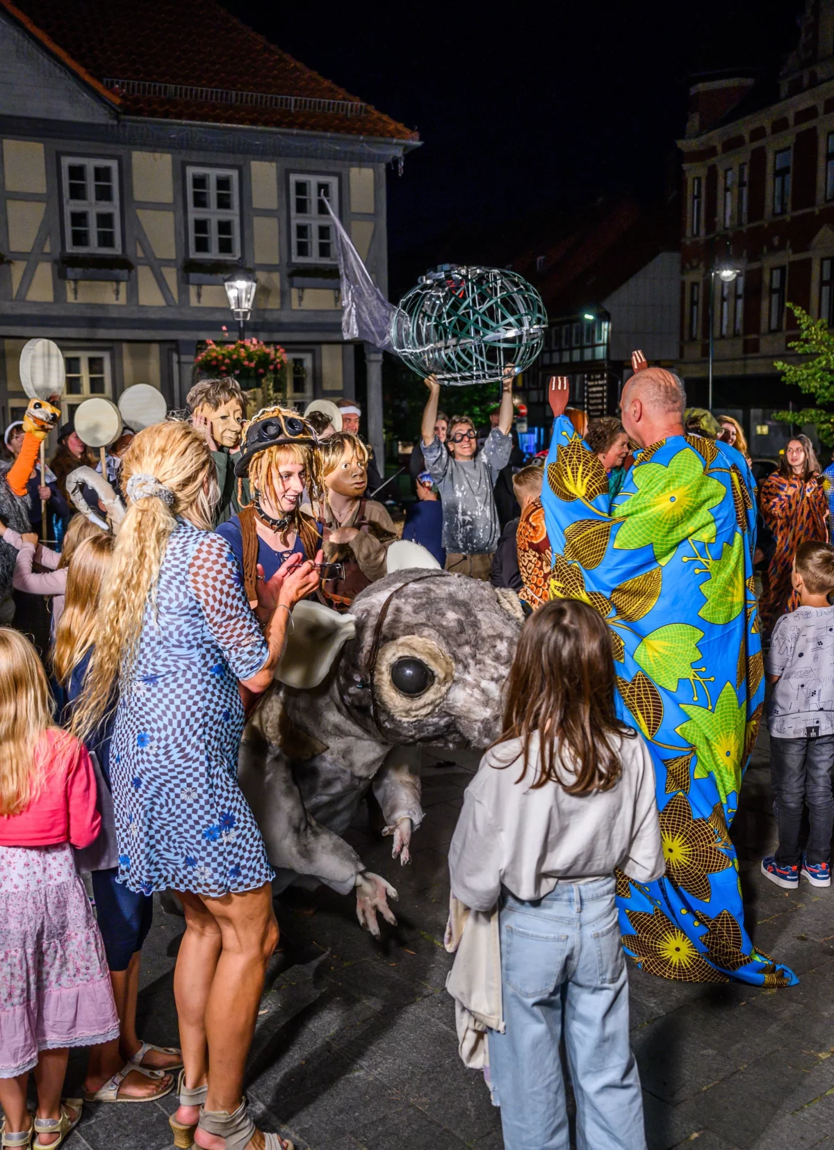 Marketplace. A crowd of adults and children. In between, people in colorful robes and masks. In focus a performer who is in a big rat costume. It looks like she is riding the rat.