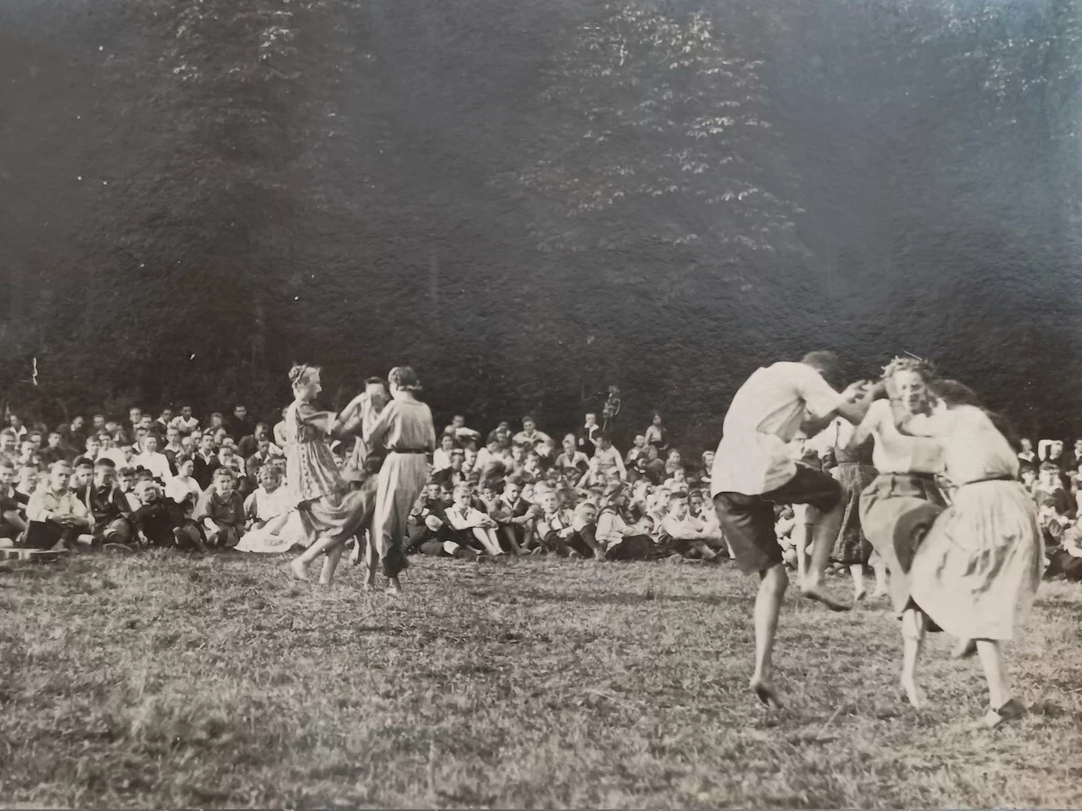 A historical black and white photograph. On a meadow, three groups of three are dancing lively in the foreground. In the background many people sit on the grass and watch.