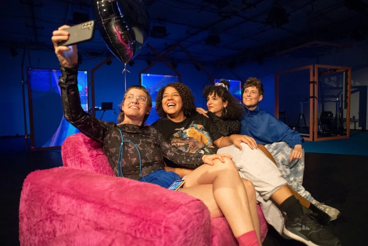 Four women are sitting on a pink plush sofa. The woman on the far left holds up a cell phone to take a selfie. They are all laughing into the camera.