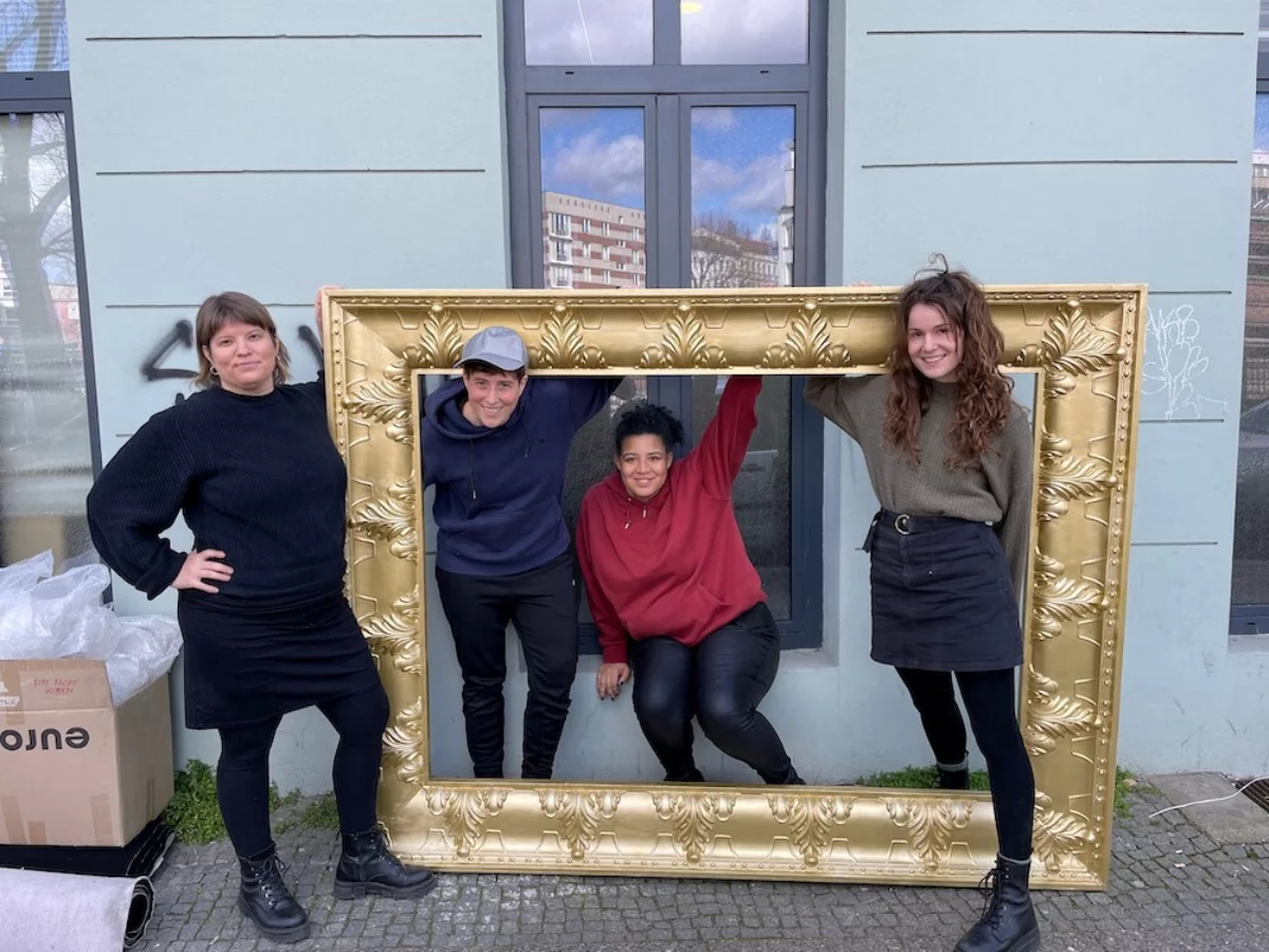 Four women pose in a large golden picture frame on the street in front of a house.