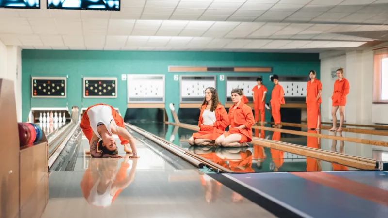 A bowling alley. On one lane a performer is dancing. On the lane next to it sit two performers, behind them stand four more performers. Everyone is watching the dancing performer.