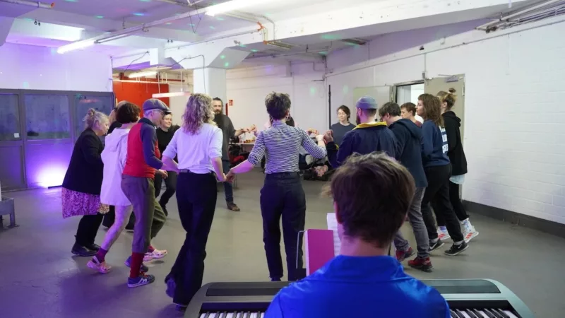 In a rehearsal room, workshop participants try their hand at a crescendo dance. In the foreground, a person sits at a piano.