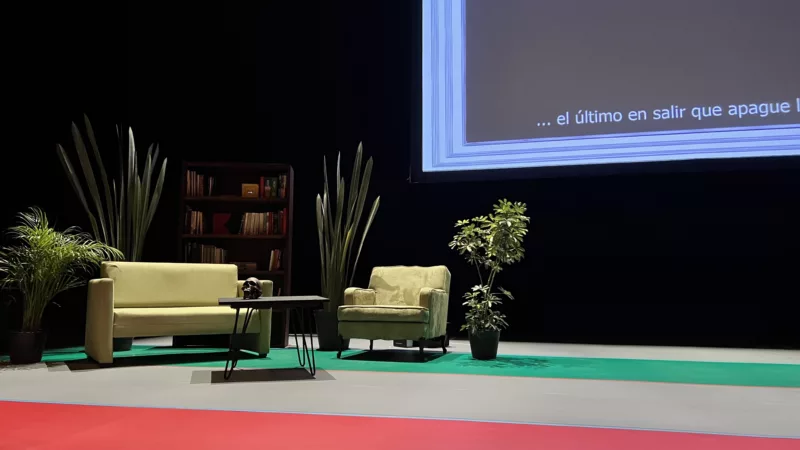 There is a sofa, an armchair and a coffee table on a stage. Behind them are a bookshelf and several large houseplants. A projection screen can be recognized in the background.
