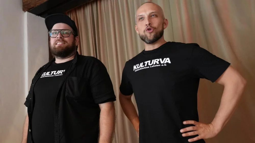Die beiden Schauspieler Sandro Šutalo und Christian Bojidar Müller stehen vor einem beigen Vorhang. Beide tragen ein schwarzes T-Shirt mit weißen "Kulturva"-Schriftzug