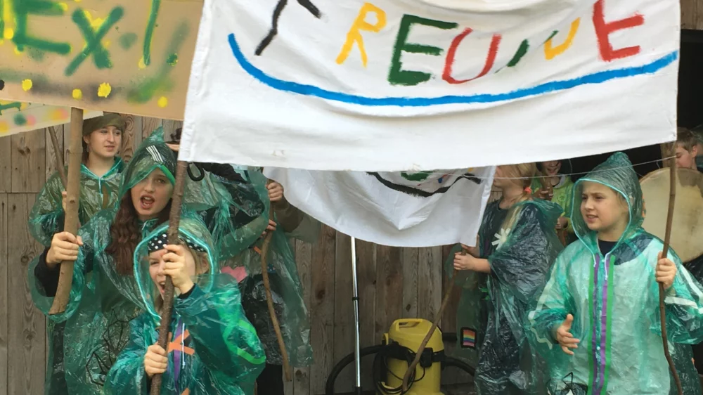 A group of children in green rain ponchos made of thin plastic demonstrate, two children hold a banner with the word Friends written on it in colorful letters.